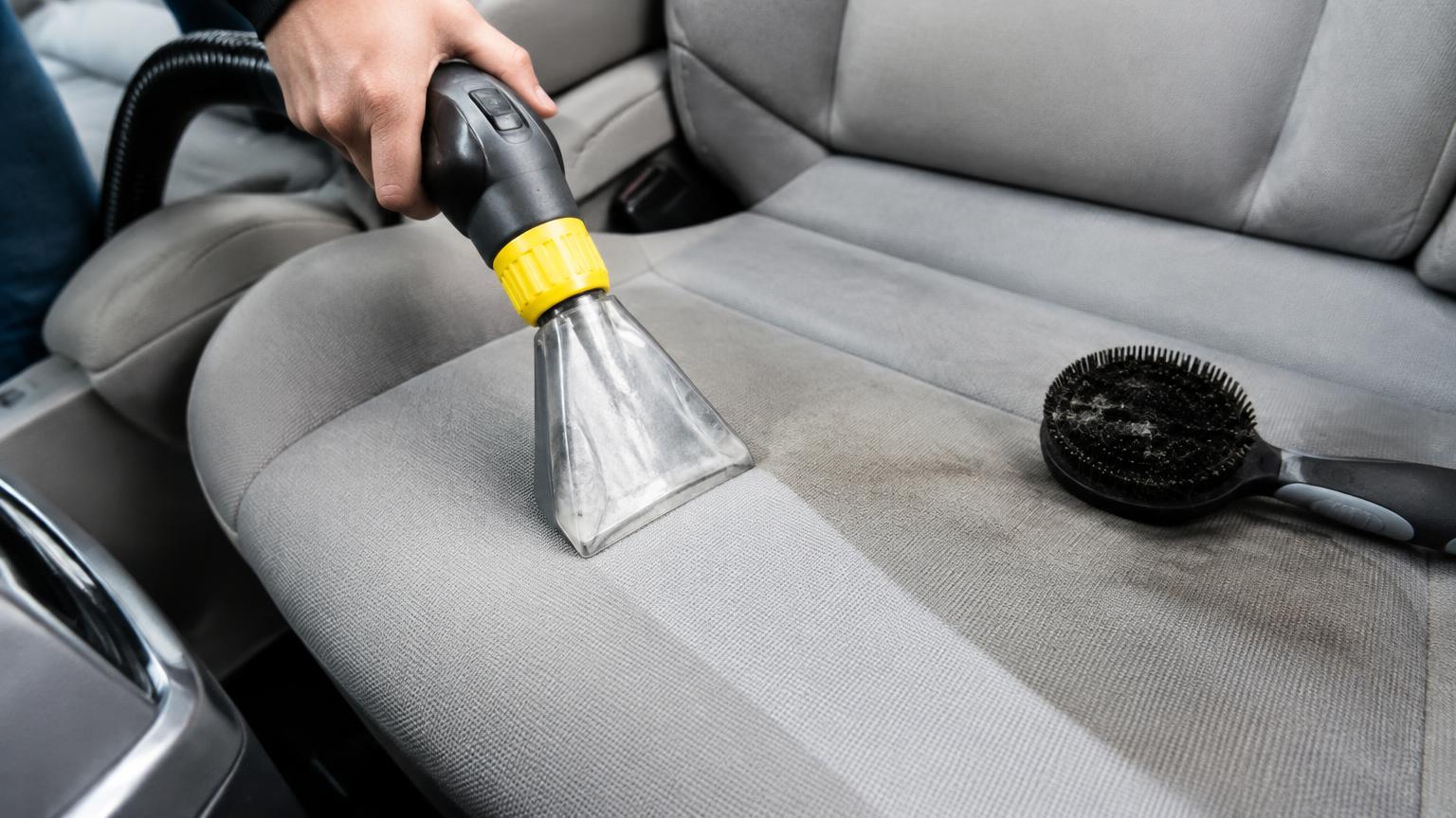Hot-water extractor cleaning a clean stripe through a stain on a grey cloth car seat, with a pet hair brush beside it