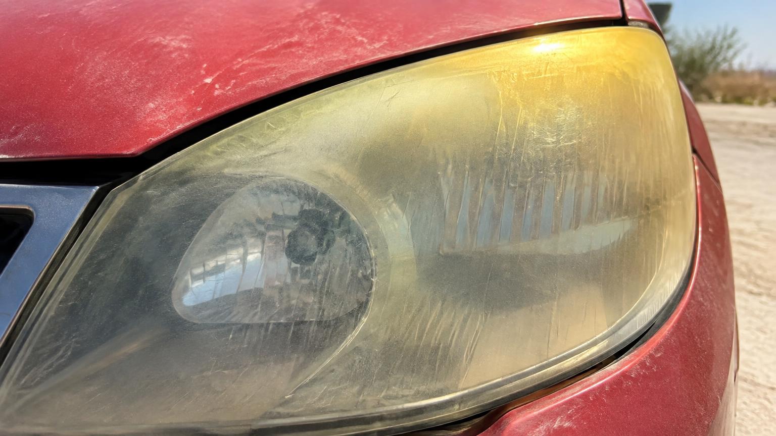Close-up of a yellowed, oxidized headlight lens with the heaviest yellowing along the sun-facing top edge, next to faded red paint
